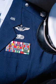 A formal military uniform featuring a collection of service ribbons, pins, and insignia on a navy blue coat. Visible is a white peaked cap with a black visor beside it. The uniform displays symbols of rank and recognition.