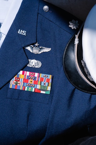 A formal military uniform featuring a collection of service ribbons, pins, and insignia on a navy blue coat. Visible is a white peaked cap with a black visor beside it. The uniform displays symbols of rank and recognition.