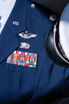 A formal military uniform featuring a collection of service ribbons, pins, and insignia on a navy blue coat. Visible is a white peaked cap with a black visor beside it. The uniform displays symbols of rank and recognition.