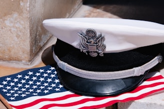 A white naval officer's cap with a black visor and silver insignia rests on a United States flag, which features white stars on a blue background and red and white stripes.