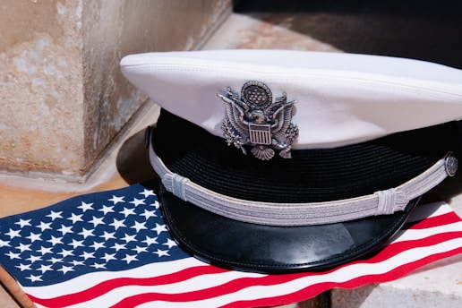 A white naval officer's cap with a black visor and silver insignia rests on a United States flag, which features white stars on a blue background and red and white stripes.