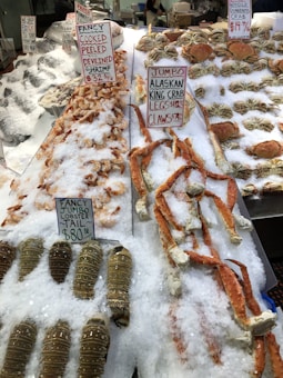 A display of various seafood items on ice at a market, including cooked peeled deveined shrimp, jumbo Alaskan king crab legs, claws, jumbo lobster tails, and whole Dungeness crabs. Each item is labeled with price tags and descriptions are written on white boards in red marker. The seafood is arranged neatly on beds of crushed ice.