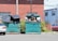 A pair of large green dumpsters are situated in a parking lot area next to a brick building. The dumpsters are partially open, with one filled with black trash bags and various discarded items, such as cardboard and plastic crates. A silver car is parked nearby, and there's an industrial building in the background. The area is surrounded by some greenery and a utility pole.