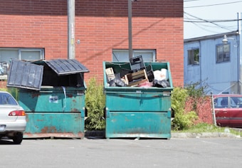 A pair of large green dumpsters are situated in a parking lot area next to a brick building. The dumpsters are partially open, with one filled with black trash bags and various discarded items, such as cardboard and plastic crates. A silver car is parked nearby, and there's an industrial building in the background. The area is surrounded by some greenery and a utility pole.