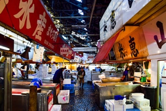 A bustling indoor fish market with bright, colorful signage displaying Japanese characters. Several vendors and workers are present, some focused on their tasks while others interact with customers. The market is filled with various stalls, boxes, and containers. The ceiling is high, with metal beams visible, adding an industrial feel to the space.