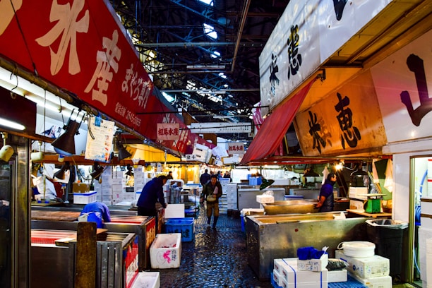 A bustling indoor fish market with bright, colorful signage displaying Japanese characters. Several vendors and workers are present, some focused on their tasks while others interact with customers. The market is filled with various stalls, boxes, and containers. The ceiling is high, with metal beams visible, adding an industrial feel to the space.