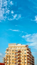 A multi-story residential building with numerous balconies and apartments is shown under a bright blue sky with scattered white clouds. The building has a combination of beige and light brown colors with some orange elements.