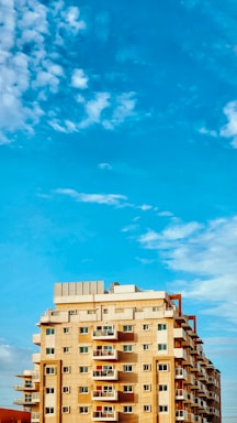 A multi-story residential building with numerous balconies and apartments is shown under a bright blue sky with scattered white clouds. The building has a combination of beige and light brown colors with some orange elements.
