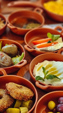Various earthenware dishes filled with an assortment of Middle Eastern or Mediterranean foods including sliced tomatoes and cucumbers, falafel, creamy dips with herbs, olives, and pickles. Each dish is arranged attractively with garnishes of mint and other herbs.