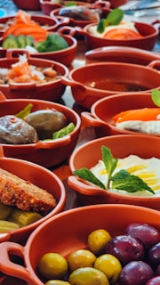 A collection of terracotta dishes filled with a variety of Mediterranean appetizers and sides. The spread includes vibrant olives, dips garnished with fresh mint, sliced cucumbers, carrots, and other vegetables. The arrangement is colorful and appetizing, emphasizing fresh ingredients and a rich assortment of textures.