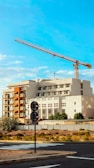 A large construction crane towers over a multi-story building with a modern design. The structure features a mix of white and earthy tones, with a prominent section painted in orange. The foreground includes a road, some sparse vegetation, and traffic signs. The sky is clear with few clouds, indicating a sunny day.