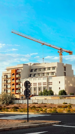 A large construction crane towers over a multi-story building with a modern design. The structure features a mix of white and earthy tones, with a prominent section painted in orange. The foreground includes a road, some sparse vegetation, and traffic signs. The sky is clear with few clouds, indicating a sunny day.