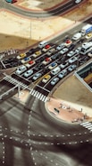 An aerial view of a busy traffic intersection with multiple lanes filled with vehicles such as cars, vans, and taxis. Pedestrians are walking on crosswalks and sidewalks. The scene is surrounded by open sandy areas and modern infrastructure with clear road markings.