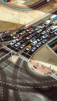 An aerial view of a busy traffic intersection with multiple lanes filled with vehicles such as cars, vans, and taxis. Pedestrians are walking on crosswalks and sidewalks. The scene is surrounded by open sandy areas and modern infrastructure with clear road markings.