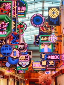 A lively and colorful indoor space filled with a variety of neon signs featuring different shapes, symbols, and text in multiple languages. The signs are suspended from the ceiling against a backdrop of a glass roof, creating a vibrant and bustling atmosphere. There is also a pink blossoming tree visible at the bottom of the scene.