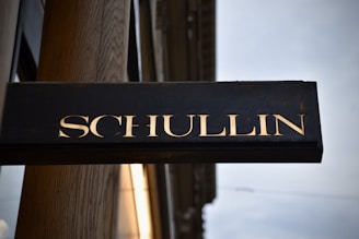 A rectangular black sign with the word 'SCHULLIN' in large, bold, gold letters. The sign is mounted on a wooden surface with a slightly cloudy sky in the background. The material of the letters appears to be shiny, possibly made of metal.