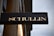 A rectangular black sign with the word 'SCHULLIN' in large, bold, gold letters. The sign is mounted on a wooden surface with a slightly cloudy sky in the background. The material of the letters appears to be shiny, possibly made of metal.