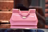 A glossy pink handbag with gold-tone hardware, featuring a small handle and a structured silhouette. It is placed on a reflective surface, with a blurred background that suggests a store display.