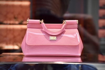 A glossy pink handbag with gold-tone hardware, featuring a small handle and a structured silhouette. It is placed on a reflective surface, with a blurred background that suggests a store display.