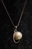 A close-up view of an elegant pearl pendant necklace, featuring a large, luminous pearl encircled by a delicate, shimmering metal setting, all set against a dark, textured background.