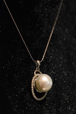 A close-up view of an elegant pearl pendant necklace, featuring a large, luminous pearl encircled by a delicate, shimmering metal setting, all set against a dark, textured background.