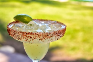 A cocktail glass filled with a chilled drink garnished with a slice of lime. The rim of the glass is coated with chili powder, and there are ice cubes within the liquid. The background shows a blurred, lush green environment, suggesting an outdoor setting.