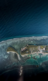 An aerial view of an island, surrounded by deep blue ocean waters and bordered by lighter, shallow areas indicating reefs or sandbars. The island is densely covered with greenery and has visible structures, including roads and buildings, laid out in an orderly fashion. The coastline features a small pier extending into the water.