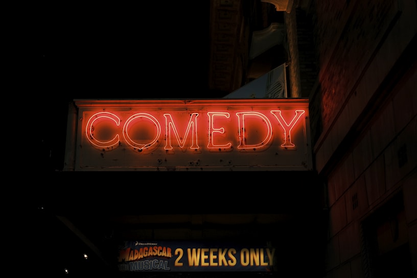 A neon sign displaying the word 'COMEDY' glows brightly against a dark background. Below this, a banner advertises 'Madagascar the Musical' with the words '2 Weeks Only' highlighted. The scene suggests a theater or comedy club setting at night.
