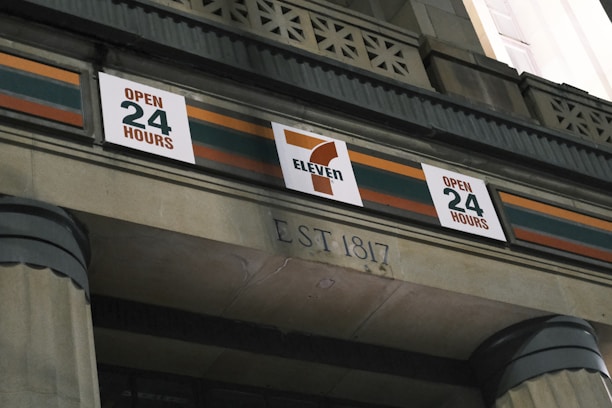 The image features the facade of a building with a 7-Eleven sign prominently displayed, indicating that the store is open 24 hours. The architectural style includes decorative elements and pillars, with the establishment year inscribed below.