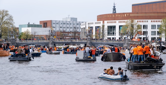A bustling river scene with numerous boats populated by people wearing orange clothing, indicating a festive event. The backdrop features a modern building labeled 'Opera & Ballet,' and there are trees and a bridge with spectators gathered along the sides.