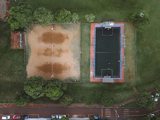 An aerial view of two adjacent sports fields, one with sand, likely for beach volleyball, and the other a synthetic turf field, possibly for soccer. Surrounding the fields are green trees and a small structure with a tiled roof. There are a few people on both fields engaged in casual play. Various parked cars are visible at the bottom, next to a paved road.