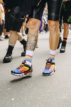 People are walking on a street wearing colorful clothing, including rainbow-striped socks and black shorts. The focus is on vibrant sneakers with a blend of colors like purple, blue, and orange. The setting appears to be a casual outdoor gathering or event.