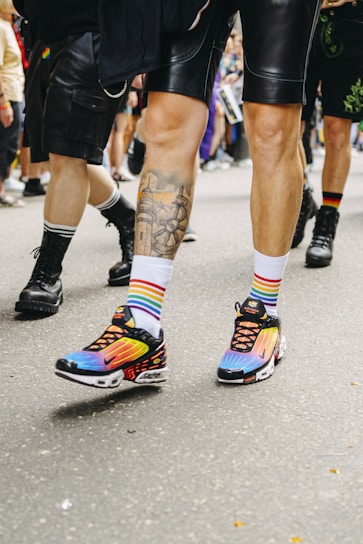 People are walking on a street wearing colorful clothing, including rainbow-striped socks and black shorts. The focus is on vibrant sneakers with a blend of colors like purple, blue, and orange. The setting appears to be a casual outdoor gathering or event.