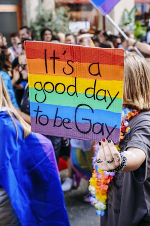 A person is holding a sign with a rainbow background stating 'It's a good day to be Gay' during a parade or rally. The scene is crowded with people, some wrapped in colorful flags, creating a vibrant and celebratory atmosphere.