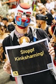 A person wearing a colorful dog mask and a necklace is holding a sign with the text 'Stoppt Kinkshaming!'. The background shows a crowd of people, some dressed in costumes or unique clothing, gathered at an event.