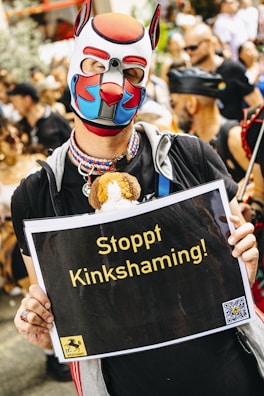 A person wearing a colorful dog mask and a necklace is holding a sign with the text 'Stoppt Kinkshaming!'. The background shows a crowd of people, some dressed in costumes or unique clothing, gathered at an event.