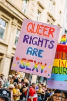A vibrant protest sign reading 'Queer Rights Are Sexy' is held up in a crowd, with colorful and bold lettering. The background is filled with people, some wearing rainbow-themed outfits, indicating a pride or LGBTQ+ event. Nearby, another sign displays rainbow colors.