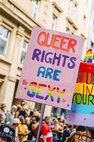 A vibrant protest sign reading 'Queer Rights Are Sexy' is held up in a crowd, with colorful and bold lettering. The background is filled with people, some wearing rainbow-themed outfits, indicating a pride or LGBTQ+ event. Nearby, another sign displays rainbow colors.