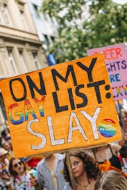 A vibrant protest scene featuring people holding signs advocating for LGBTQ+ rights. The focal point is a large orange sign with the text 'On My Gay List: Slay' in bold letters. The crowd is colorful and engaged, with participants displaying symbols of pride, such as rainbow flags.