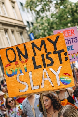 A vibrant protest scene featuring people holding signs advocating for LGBTQ+ rights. The focal point is a large orange sign with the text 'On My Gay List: Slay' in bold letters. The crowd is colorful and engaged, with participants displaying symbols of pride, such as rainbow flags.