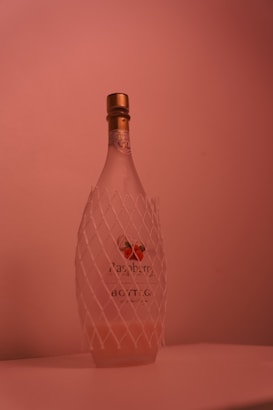 A frosted glass bottle with a golden cap stands upright. The bottle features a label with images of raspberries and has a protective white netting wrapped around it. The lighting creates a warm, soft pink hue over the entire scene.