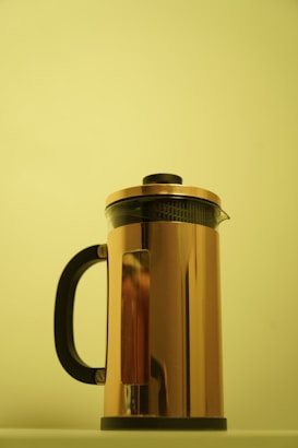 A metallic French press with a gold reflective surface stands against a plain light yellow background. The design includes a black handle and lid, creating a simple yet elegant household object.
