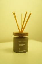A reed diffuser with wooden sticks is placed in an elegant, matte-finished container labeled 'Neon Sheep' and 'mahogany vetiver.' The background and lighting have a warm, yellow tint, giving the scene a cozy appearance. The container sits on a light-colored surface, with the wood lid complementing the natural aesthetic.