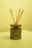 A reed diffuser with wooden sticks is placed in an elegant, matte-finished container labeled 'Neon Sheep' and 'mahogany vetiver.' The background and lighting have a warm, yellow tint, giving the scene a cozy appearance. The container sits on a light-colored surface, with the wood lid complementing the natural aesthetic.