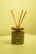 A reed diffuser with wooden sticks is placed in an elegant, matte-finished container labeled 'Neon Sheep' and 'mahogany vetiver.' The background and lighting have a warm, yellow tint, giving the scene a cozy appearance. The container sits on a light-colored surface, with the wood lid complementing the natural aesthetic.