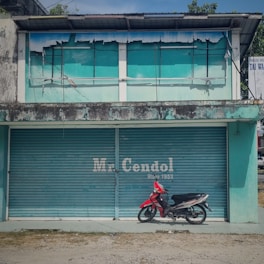 An old, teal-colored building with peeling paint and a rusty exterior. The shutters are closed with 'Mr. Cendol Since 1952' painted on them. A red motorcycle is parked in front of the building.