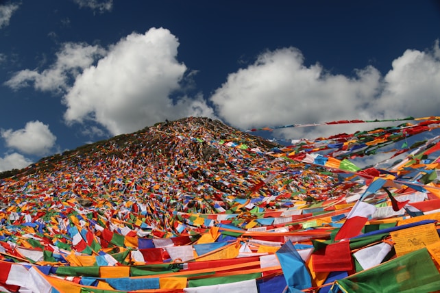 A hillside covered with an extensive display of colorful prayer flags flutters under a bright blue sky. The vibrant flags, in shades of red, yellow, blue, green, and white, are strung across the landscape, creating a dynamic and spiritual scene. The background features soft, white clouds scattered across a deep blue atmosphere.