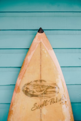 A vintage surfboard with a pointed tip is propped against a turquoise wooden wall. The surfboard has a worn appearance with a design featuring a wave and palm trees, along with the text 'Swell Patrol'.