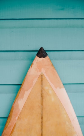A surfboard with a pointed tip rests against a backdrop of teal wooden slats. The surfboard features an aged, yellowish hue with black detailing at the tip.