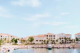 A picturesque marina bordered by Mediterranean-style buildings with terracotta roofs. Several boats are docked along the calm waterfront. The scene is enhanced by clear blue skies and lush green palm trees lining the area.